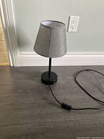 Side view image of the black touch lamp with grey lampshade placed on gray wood floor, showing cord and inline switch