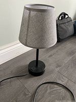 Touch lamp with black base and grey fabric shade placed on wood floor near wall baseboard.