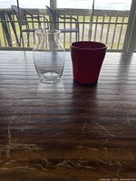 Photo showing a clear glass vase and a red plant pot on a dark wooden table with a window background.