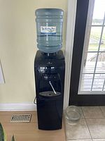 Full view of a tall black water cooler with a 5-gallon water bottle on top against a wall with a window and door visible. A clear glass bowl sits on the floor to the right of the cooler.