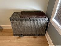 View of the grey synthetic wicker laundry basket with lid and two metal foot pedals for opening, placed on a wooden floor.