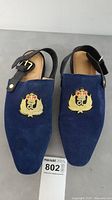 Pair of navy blue suede Christian Louboutin slingback flats side by side showing embroidered crest and black strap with gold buckle