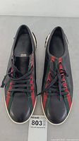 Top front view of both black Gucci sneakers with green and red web stripe along lace area and black laces.