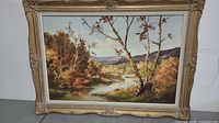 Full view of the framed painting showing autumn landscape with a stream and trees.