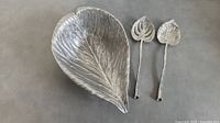 Leaf-shaped metal salad bowl with two matching leaf-shaped serving spoons on a table.
