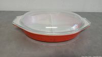 Side angled view of the red divided Pyrex casserole dish with clear glass lid on top, showing its oval shape and integrated handles.