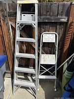 Two aluminum ladders side by side: a 6 ft step ladder and a smaller folding step stool against a wooden fence