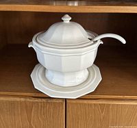 Soup tureen with lid, ladle resting in lid notch, and octagonal underplate on a wooden shelf.