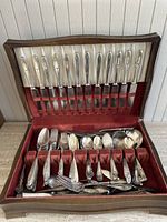 Photo of the full silverware set inside a wooden case with red and white lining, showing knives slotted in the lid and various forks, spoons, and serving utensils arranged in the bottom section.