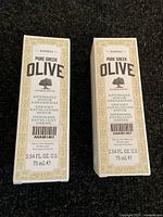 Front view of two boxed Korres Pure Greek Olive Creamy Exfoliating Scrub 75ml tubes side by side on a dark surface.