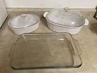 Three casserole and baking dishes: two white stoneware oval casseroles with clear glass lids, one rectangular clear glass baking dish without lid