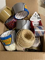 Box containing assorted tape rolls including duct tape, packing tape, Scotch tape, and twine spools