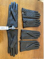 Three pairs of black faux leather gloves laid flat on wooden surface, showing the plain pair with tags and the two quilted pairs with bow embellishments