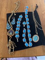 Top-down view of jewelry set on black cloth, showing gold-tone necklace with tassel pendant, two silver-tone blue stone bracelets, silver-tone blue stone pendant necklace, and blue stone earrings.