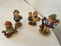 Photo of four Hummel porcelain figurines grouped together showing a boy sitting on a tree branch, a girl with a red hat, a standing boy, and two children holding baskets.