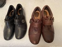 Two pairs of Clarks Artisan leather loafers, one black and one brown, size 9, showing top view and Velcro strap with button accent.