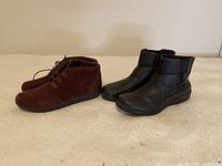 Two pairs of ankle boots side by side on a light surface, one black leather with zip closure and one burgundy suede with lace-up closure.