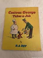 Front cover of Curious George Takes a Job book showing yellow background, title, and illustration of Curious George with a baker and mailman