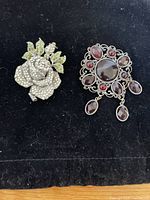 Two decorative brooches displayed on black fabric, showing front view of each piece with rhinestone details.
