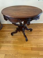 Full view of oval dark brown wooden accent table with carved legs and pedestal base on hardwood floor.