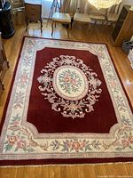 Full view of rectangular handmade wool floral area rug showing red central field and beige floral borders with floral medallion center.
