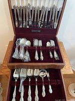 Full view of the Heritage flatware set displayed in an open wooden case with burgundy velvet lining. Includes all utensils arranged neatly showing the set's organization.