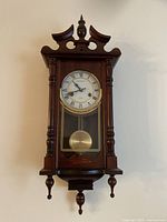 Full front view of the Kassel wooden wall clock showing the decorative wooden top and pendulum section.