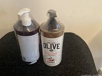 Two pump bottles of Korres Pure Greek Olive bath products, one with white pump and one with black pump, sitting on fabric surface.