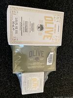 Three Korres Pure Greek Olive skin care products stacked; Vitality Shield Booster Oil box on top, soap bar below, and a small cream box.