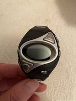 Close-up photo of the black Mio Shape fitness watch showing the oval digital display and three control buttons around the screen.