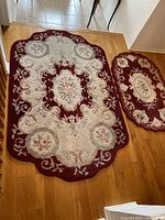 Large and medium oval wool rugs with rippled edges, showing detailed floral patterns in red and cream tones on wood floor.
