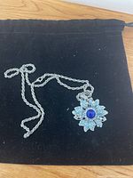 Silver-toned necklace with medium-sized flower-shaped blue crystal pendant on black cloth background.