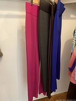 Four pairs of Nygard Slims women's pants hanging on wooden hangers, visible colors magenta, gray, brown, and royal blue.