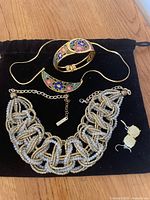 Photo showing the gold tone and white beaded statement necklace, floral inlay bracelet, necklace, and yellow square earrings all displayed on a black fabric surface.