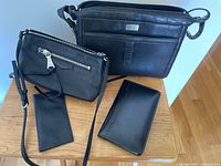Four items displayed on wooden surface: two handbags, a cheque book cover, and a photo book all in black leather or faux leather.