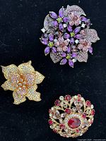 Three floral brooches displayed on black background showing designs and rhinestones.