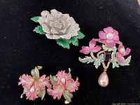 Top view of lot showing three jewelry pieces on black velvet. Includes the featured brooch with pink flowers, silver glitter floral brooch, and a pair of pink earrings.