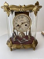 Front view of the clock showing gold-toned metal frame with ornate top and base, Roman numeral dial with visible winding holes, and clear glass enclosure.