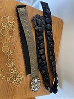 Three belts laid on wooden surface: gold-tone chain link belt, wide gold-tone metal belt with floral rhinestone buckle, and black fabric belt with large black stone embellishments and decorative clasp.