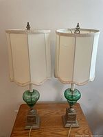 Pair of vintage green and bronze Depression glass lamps with off-white original fabric shades, one shade torn, placed on wooden table against white background.