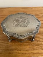 Closed zinc keepsake box with scalloped edge and engraved coat of arms on lid, resting on lion claw feet with lion faces on the front.