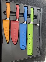 Five multi-colored Wolfgang Puck cutting utensils with plastic handles and matching sheaths in black tray.