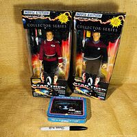 Two Star Trek Movie Edition action figures still in original packaging, standing upright with their accessories visible inside the box, accompanied by a rectangular Star Trek collectible tin placed in front.