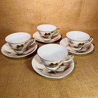 Four handpainted Japanese porcelain cups and matching saucers arranged on a woven beige fabric background, showing village scenes and mountain motifs in brown and pink on white.