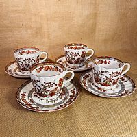 Four porcelain cups and saucers set with brown/orange floral and gold detailed India Tree pattern arranged on woven textured background.