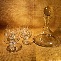 Photo showing one round clear glass decanter with stopper and four short stemmed glasses in the background on a textured brown surface.