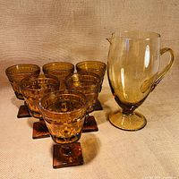 Overall lot view showing six goblet-style glasses and one pitcher in amber glass placed on a fabric surface.