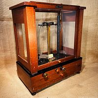 Front and right angle view of the antique scale inside a wooden and glass case, showing the mechanism and wooden drawer closed.