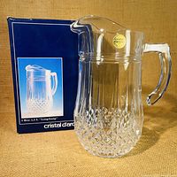 Photo of the Cristal d’Arques lead crystal pitcher standing next to its original blue box with product image and labeling.