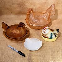 Four hens on nests displayed on burlap background with marker for scale, showing pink depression glass, amber glass, ceramic salt and pepper shakers, and milk glass.